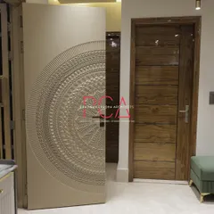 The bespoke main door of 'The Cursive Abode', shown here slightly ajar. The intricate exterior carving contrasts with the clean, modern wooden door of the adjacent room, highlighting a blend of styles.