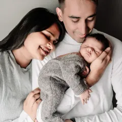 A classic and intimate studio portrait of new parents holding their newborn baby. The simple grey background and coordinated white outfits keep the focus on the family's loving bond.