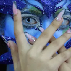 A striking close-up of a fantasy makeup look. The use of contact lenses, detailed linework, and custom nail art transforms the model into a creature from another world.