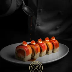 The final touch. A chef carefully drizzles a finishing sauce on a row of exquisite salmon and tuna sushi rolls, each topped with vibrant tobiko. This is the artistry and attention to detail we pour into every single dish.