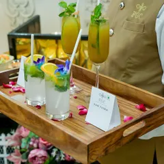 A server presents a tray of refreshing mocktails, including Aam Panna. The drinks are garnished with fresh mint, fruit slices, and edible flowers, demonstrating our commitment to beautiful presentation in every detail.