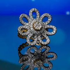 A close-up of a diamond-studded, flower-shaped ring set against a vibrant blue background with its reflection. This macro jewellery shot highlights the sparkle of the stones and the precise detail of the setting.