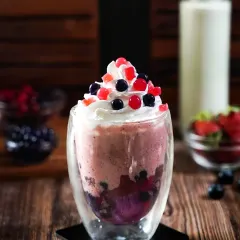 A layered berry boba smoothie in a double-walled glass, topped with whipped cream and colorful popping boba. The dark, rustic setting makes the vibrant colors of the drink stand out.