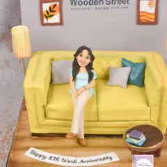 A 10th work anniversary cake for an employee at a furniture company. The cake is carved into the shape of a sofa, complete with a custom figurine, cushions, and a miniature living room scene.
