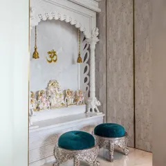 An elegant, all-white marble puja room from our 'Urban Oasis' project. The mandir features intricate carvings, golden 'Om' accents, and two plush turquoise stools for seating.