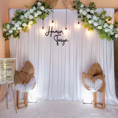 A minimalist and modern wedding backdrop. This elegant setup features a clean white curtain, simple hanging bulbs, and delicate white floral arrangements, perfect for a chic and contemporary ceremony.