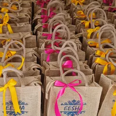 We create bespoke return favours like these custom-printed jute bags for a wedding. The bags feature the bride's name and wedding date, with vibrant ribbons in yellow and pink.