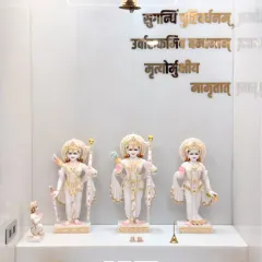 A close-up of the beautiful marble idols of Ram, Sita, and Lakshman, set against a pristine white backdrop with a hanging brass bell, creating a pure and peaceful focal point.