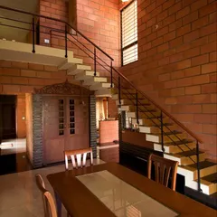 At the Sharada Residence, an exposed brick wall provides a dramatic backdrop for the dining area and floating staircase. The design integrates a traditionally carved wooden door, blending rustic materials with classic Indian craftsmanship.