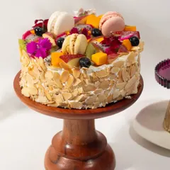 A vibrant fresh fruit cake, generously topped with a variety of seasonal fruits like mango, kiwi, and dragon fruit, plus colorful macarons.
