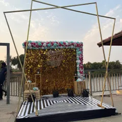 We can create a complete, stylish party stage for outdoor events. This setup features a gold sequin backdrop under a geometric metal arch, with a custom 'Party' neon sign and a patterned floor, perfect for a landmark birthday celebration by the water.