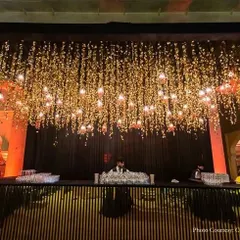 This modern bar counter features a black slatted wood design. The space above is decorated with a stunning installation of hanging flowers and lights, creating a dramatic and sophisticated look for an evening event.