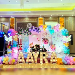 A vibrant fairy and butterfly themed first birthday stage for Aayra. The decor features marquee letters, a sequin backdrop, colorful balloon garlands, and cute fairy cutouts, all coming together to create a cheerful and magical setting.