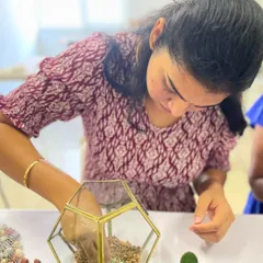 A participant carefully places the first layer of gravel in her geometric terrarium, the crucial foundation for proper drainage.