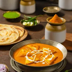 Shahi Paneer in a traditional metal bowl, surrounded by pickling jars and parathas. This composition for Rakesh Masala creates a feeling of a complete, wholesome meal.