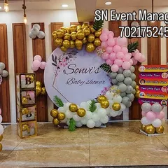 Here is an elegant baby shower setup for a party hall. It features a personalized circular backdrop with the mother's name, surrounded by a custom balloon arrangement in pink, gold, and sage green. I can match any color scheme you have in mind.