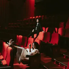A stylish, full-length shot of the couple owning the theatre space. Her relaxed pose with feet up and his confident stance on the seat back create a playful, high-fashion image that tells a story of a modern power couple.
