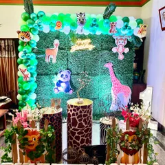 A jungle-themed decoration perfect for a home setup. It features a green hedge backdrop adorned with animal cutouts, animal-print plinths, and marquee letters for the birthday boy's name.