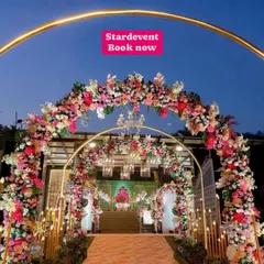 A luxurious wedding entrance at dusk. The walkway is lined with multiple floral arches in pink and white, illuminated by warm lights and golden pillars, creating a magical tunnel effect.