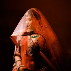 A stunning bridal portrait with dramatic lighting. The bride is veiled and looks down, creating a sense of serenity and grace. The rich red tones make this a powerful and artistic image.