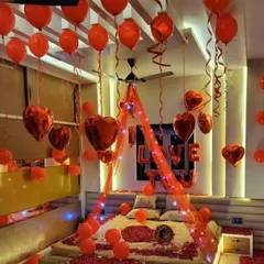 A passionate and romantic room setup entirely in red. The bed is decorated with a canopy, fairy lights, and rose petals, while red heart-shaped and latex balloons fill the room, creating an atmosphere of pure love.