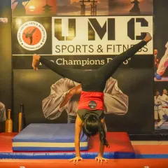 A gymnast holds a perfect handstand split on the balance beam, a move that requires immense strength and balance. Our UMC logo in the background represents the supportive environment where such skills are honed.