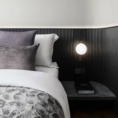 This custom headboard features fine black fluted paneling and an integrated stone side table. A soft, ambient light strip at the top and a modern wall sconce provide layered lighting for a cozy atmosphere.