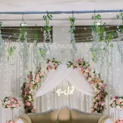 A dreamy Nikah backdrop with a floral arch and draped canopy. The decor features hanging wisteria and a 'Qubool Hai' neon sign, creating a soft and romantic ambiance.