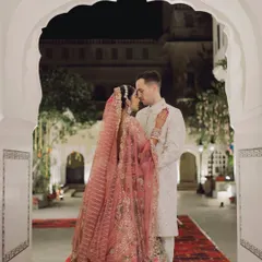 A romantic couple portrait framed by a classic scalloped archway. The bride's intricate pink lehenga and the groom's elegant sherwani are beautifully captured in this timeless shot.