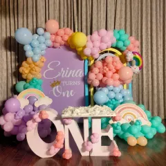 Even a small space at home can be magical. Here is a pastel rainbow theme for a first birthday, featuring a custom backdrop for "Erina turns One" and large "ONE" marquee letters decorated with matching balloons.
