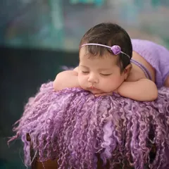 A newborn girl in a simple purple outfit rests her head on her hands, sleeping on a fluffy purple textured blanket.