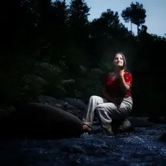A dramatic portrait of a woman sitting by a river at dusk. The creative lighting highlights her against the dark, natural surroundings, creating a moody and contemplative lifestyle photograph.