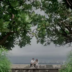 A serene, wide shot of a couple sitting at Marine Drive, framed by lush green trees. It captures a peaceful moment amidst the bustling city.
