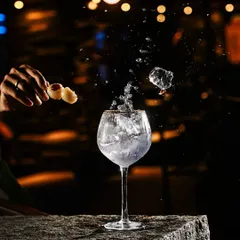 A dramatic action shot of a mixologist creating "The Panic Drink." Ice splashes into a glass filled with a lavender-hued concoction, garnished with lychees, showcasing the artistry and excitement of our cocktail program.