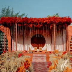A modern yet traditional mandap design for an outdoor wedding. The structure features a dramatic canopy of red florals and hanging strings of beads, set against a circular floral backdrop for a powerful visual statement.
