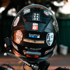 A detailed shot of a rider's helmet, covered in stickers that tell a story of their identity and community. This image focuses on the personal details that define the biker lifestyle, from GoPro mounts to group logos.
