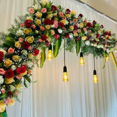 A close-up of a floral arch for an engagement stage. The arch is decorated with red and yellow roses, with hanging Edison bulbs adding a touch of modern charm.