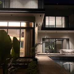 The pool deck at "The One" is softly illuminated at night, creating a serene and luxurious atmosphere. The lighting reflects off the water and highlights the large indoor living spaces, showcasing the seamless connection between the home and its landscape.