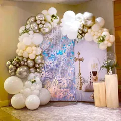 A modern First Holy Communion backdrop combining a silver shimmer wall with a white and silver balloon arch. The setup includes pedestals for cake and decor, with religious symbols incorporated into the design.