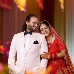 A classic wedding portrait of the couple. The bride in her traditional red lehenga rests her head on the groom's shoulder, who is dressed in a sharp white tuxedo, creating a beautiful contrast.