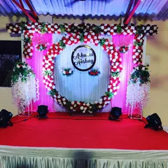 A beautiful stage for an engagement or reception, featuring a circular arch decorated with red and white flowers. A custom sign with the couple's names, 'Ashu and Akshay', is placed in the center, creating a personalized focal point on the red-carpeted stage.