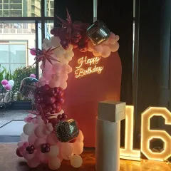 A sweet 16 birthday party with a pink arched backdrop. The decoration includes a balloon garland with disco balls and starburst accents, a neon sign, and a marquee number '16'.