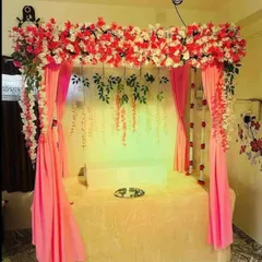 A lovely home mandap featuring a canopy of red and white flowers with flowing pink drapes. The backdrop includes hanging green vines, adding a touch of natural elegance to the festive setup.