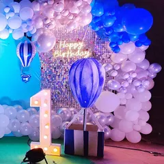 A dreamy hot air balloon theme for a first birthday. This magical setup features a large balloon basket prop, a shimmering sequin backdrop, and a cloud-like balloon garland in shades of blue and white. The light-up number '1' and neon 'Happy Birthday' sign add a warm glow.