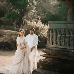 A full-length portrait of the couple standing proudly in their coordinated white ethnic outfits, set against the warm, sunlit stone of a heritage site.