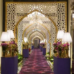 A truly grand Sufi-inspired entranceway. A series of golden arches with intricate jali screens, a rich purple carpet, and elegant floral arrangements create a long, dramatic gallery for guests to walk through.