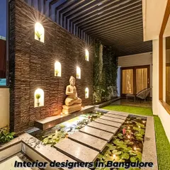 A serene and tranquil patio design featuring a stone accent wall with illuminated niches. The central water feature with a Buddha statue and lily pads creates a peaceful, meditative atmosphere.