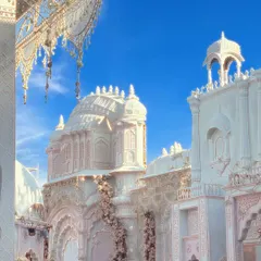 An ethereal view of a white palace structure against a brilliant blue sky. The design features delicate blush pink floral vines climbing the ornate white pillars and domes, creating a fairytale setting for a couple's special day.
