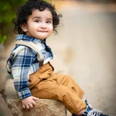 A handsome little boy with curly hair, sitting on a rock in the park. The natural setting and his stylish outfit create a timeless portrait.