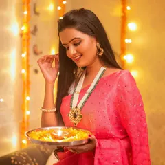 A beautiful shot of a model in a pink saree, holding a traditional puja thali. The warm lighting from the diya and fairy lights creates a magical glow.